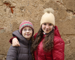 Friendship and patronage concept - two teenage girls, smaller and bigger, in winter clothes stand embracing outdoor on old grunge wall background