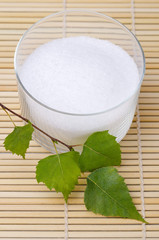 Xylitol birch sugar in a glass bowl with birch leaves on a bamboo mat. White granulated sugar alcohol, substitute used as sweetener that taste like table sugar, extracted from the wood of birch trees.