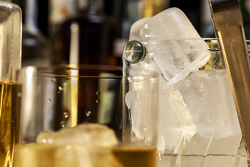 Ice cubes into a glass ice bucket seen behind a whisky cup