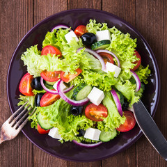 Greek salad (lettuce, tomatoes, feta cheese, cucumbers, black olives, purple onion) on dark wooden background top view. Healthy food.