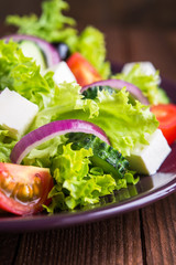 Greek salad (lettuce, tomatoes, feta cheese, cucumbers, black olives, purple onion) on dark wooden background close up. Healthy food.