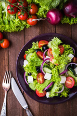 Greek salad (lettuce, tomatoes, feta cheese, cucumbers, black olives, purple onion) on dark wooden background top view. Healthy food.
