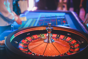 A close-up vibrant image of multicolored casino table with roulette in motion, with the hand of croupier, and a group of gambling rich wealthy people in the background
