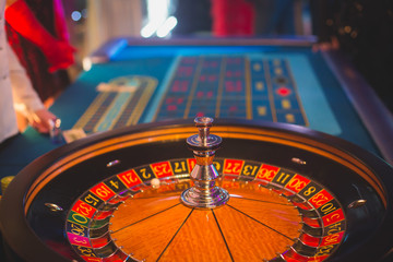 A close-up vibrant image of multicolored casino table with roulette in motion, with casino chips. the hand of croupier, mone and a group of gambling rich wealthy people in the background
