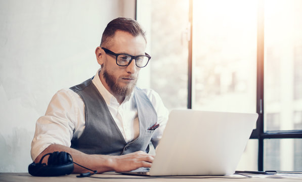 Stylish Bearded Businessman Wearing Glasses White Shirt Waistcoat Working Modern Office Startup Project.Creative Young Guy Using Laptop Computer Wood Table Workplace.Panoramic Windows Background.Flare
