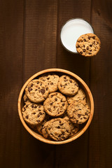 Chocolate Chip Cookies oder Kekse mit Schoko-Tröpfchen in Holzschüssel mit einem Glas Milch, fotografiert auf dunklem Holz mit natürlichem Licht