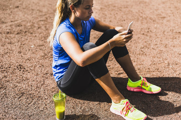 Sportive young woman sitting on sports field with cell phone and drinking mug