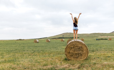 Back view of teenage girl standing on straw bale on a meadow raising arms