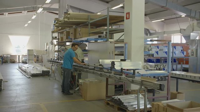 A worker in the factory is looking for some material in the table that he will later use. Wide-angle shot. There are many boxes and machines around.
