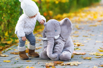The baby girl and toy elephant are in the autumn city park.
