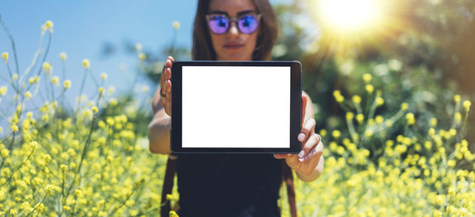 Hipster holding in hands tablet computer. Girl traveler with sunglasses using gadget on sun flare and yellow flowers background, mockup of blank empty screen copy space for text, female photographer