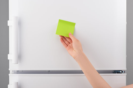 Female hand holding blank green paper note on white refrigerator