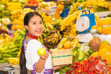 Beautiful young hispanic woman wearing andean traditional blouse posing for camera holding basket of fuits inside fruit market, colorful healthy food selection in background