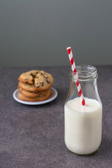 A bottle of milk with a classic red and white straw. Homemade chocolate chip cookies in the background 