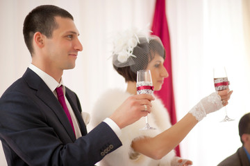 Newlyweds raise their glasses decorated with laces and red ribbo