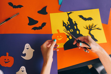 close up of female hands with scissors cut paper orange pumpkin on the background Halloween composition. Castle, ghosts, bats, pumpkins are cut from colored paper. Homemade decorations for Halloween