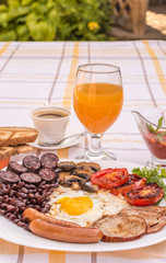 Full English fried breakfast with bacon, egg, sausages, black pudding, mushrooms, grilled tomatoes and baked beans. Closeup with toasts, cup of coffee, orange juice and home made  red sauce.