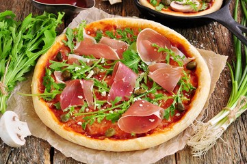 Homemade pizza with tomato sauce, mozzarella cheese, organic arugula, Parma ham, capers and parmesan cheese.