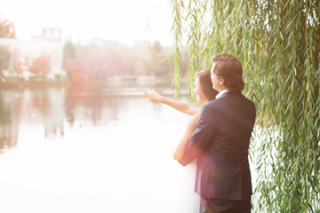 Happy wedding couple standing near the river at sunset, looking away
