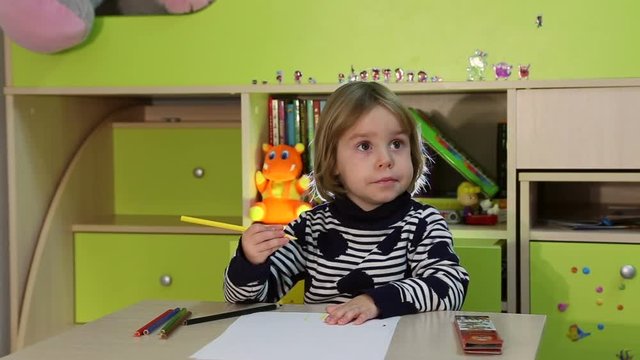 3 years old kid draws something using brush and pencil, tries to watching TV. Bookshelf on background.