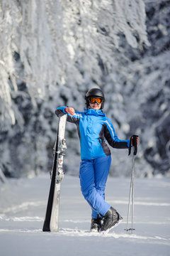 Portrait of young happy female skier standing enjoying sunny day against beautiful snow covered trees on the background. Girl is holding her skis in one hand and poles in another. Bukovel, Ukraine