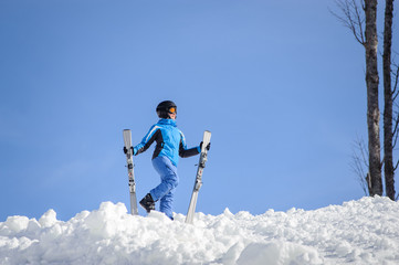 Active female skier standing on top of the mountain against blue sky on a sunny day, wearing blue ski suit helmet and goggles on sunny day. Girl is holding her skis. Winter sports concept