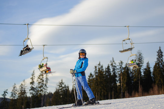 Full length portrait of young female skier against ski-lift on a sunny day. Woman is wearing helmet skiing glasses gloves and blue ski suit