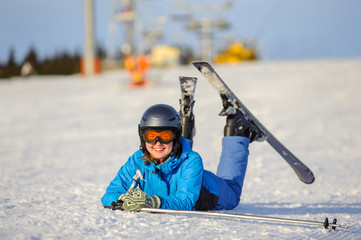 Fototapeta na wymiar Close-up portrait of young happy skier woman in blue ski suit orange goggles and helmet lying on the snow at ski resort on a sunny day. Ski vacation.