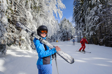 Young happy female skier on a ski slope in the winter forest on a sunny day. Woman is holding skis on her shoulder smiling and looking towards the camera. Ski resort. Bukovel, Ukraine.