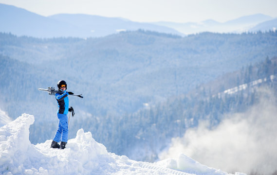 Skier standing on top of the mountain and enjoying the view on beautiful winter mountains on a sunny day. Girl is wearing blue ski suit and holding skis on her shoulder. Winter sports concept.