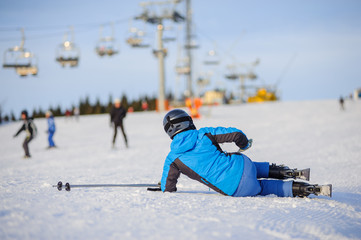 Young woman skier in blue ski suit after the fall on mountain slope trying get up against ski-lift. Ski resort. Winter sports concept.