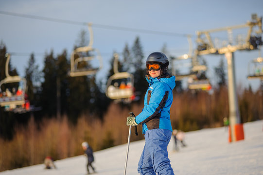 Portrait of young female skier against ski-lift in the evening. Woman is wearing helmet, skiing glasses gloves and blue ski suit