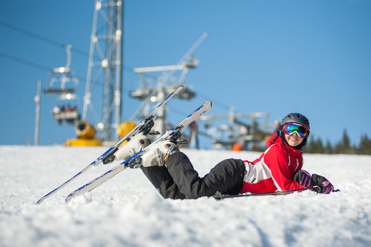 Woman smiling, lying with skis on snowy at mountain top in sunny day with ski lifts and blue sky in background.