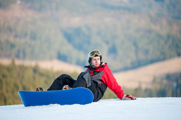 Fototapeta na wymiar Male snowboarder with snowboard wearing helmet, red jacket, gloves and pants sitting on snowy slope on top of a mountain and looking away, with an astonishing view on mountains and forest