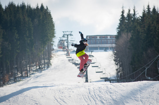 Female boarder on the snowboard jumping over the slope in winter with snowy slope and ski lifts in background, extreme sport