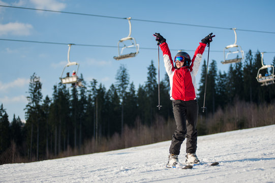 Female skier wearing helmet, red jacket and ski goggles standing on snowy slope with hands raised up in sunny day with forest and blue sky in background. Carpathian, Bukovel