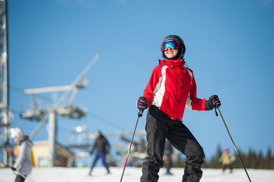 Portrait of female skier wearing helmet, red jacket and ski goggles standing with skis on mountain top at a winter resort in sunny day with ski lifts and blue sky in background.