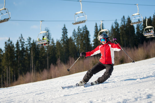 Smiling woman in the moment of falling at resort in sunny day against ski-lift, forest and blue sky. Woman is wearing red jacket, helmet and goggles. Carpathian Mountains, Bukovel