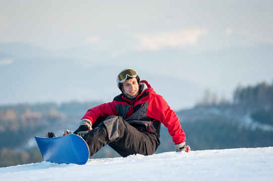 Smiling snowboarder wearing helmet, red jacket, gloves and pants sitting on snowy slope on top of a mountain, with an astonishing view on hills