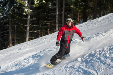 Fototapeta na wymiar Male snowboarder slides down from the mountain in winter day, overlooking the snowy slope and forest at a winter resort