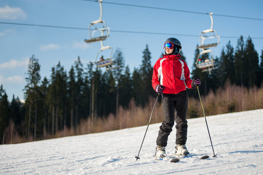 Female skier wearing helmet, red jacket and ski goggles standing with skis on snowy slope and looking away in sunny day with forest and blue sky in background.