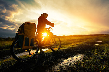 Young lady hiker with loaded bicycle standing on a wet rural road in the meadow at sunset