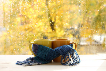 warm drinks for the autumn mood/ mugs tied together warming scarf on the background of a wet window...
