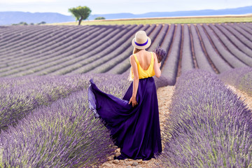 Young pretty woman in the long waving skirt and hat standing with lavender bouquet on the lavender...