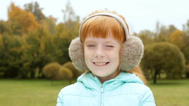 Close up cute ginger girl in the earmuffs smiling, autumn trees behind