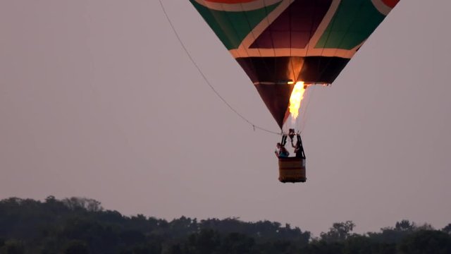 Hot air balloon in flight moving low over tree tops finding a place to land showing the burner flame heating the baloon and the pilot in basket pointing out a place to land moving steady and low 4k