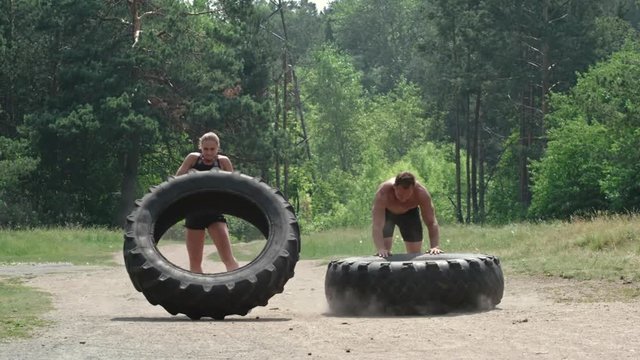 Slow motion shot of strong man and woman flipping large tractor tires towards the camera on dusty road in the park