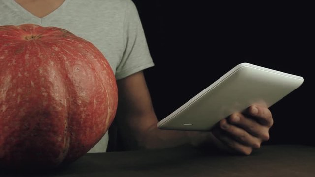 Guy in white shirt use picture on tablet computer as reference and draw on pumpkin with a marker