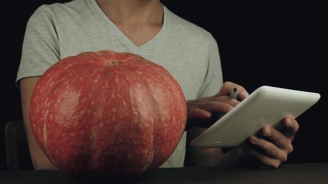 Guy in white shirt use picture on tablet computer as reference and draw on pumpkin with a marker