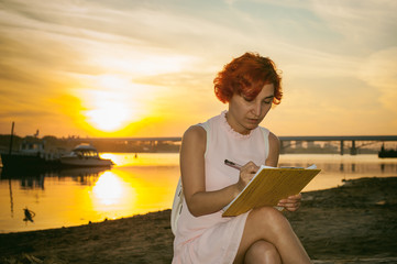 woman with dyed red hair in a pale pink dress with white backpack, signed important documents sitting outdoors near the river at sunset, against a boat mooring